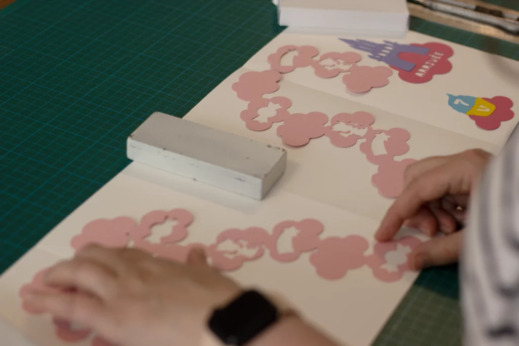 Collage des éléments décoratifs : nuages rose poudré pleins ou évidés, château, gâteau et autres formes sur le plateau de jeu "Au royaume des licornes".