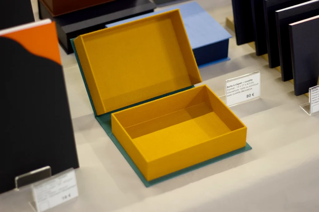 Boîte à clapet ouverte bicolore, en toile du reliure vert et jaune moutarde, avec cuvette, présentée lors du salon de l’artisanat.