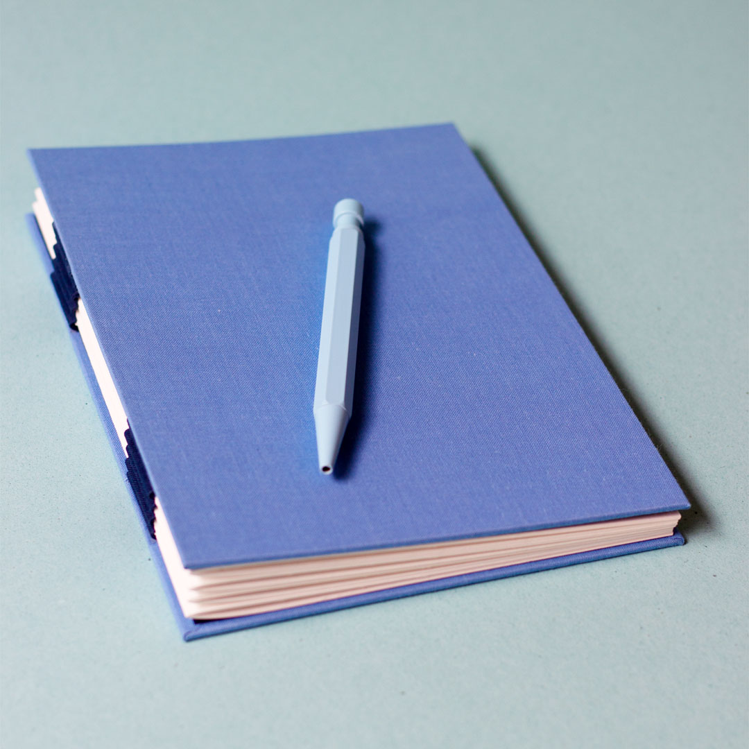 Carnet avec une reliure australienne à charnière piano inversée, style plat, accompagné d’un stylo bleu.