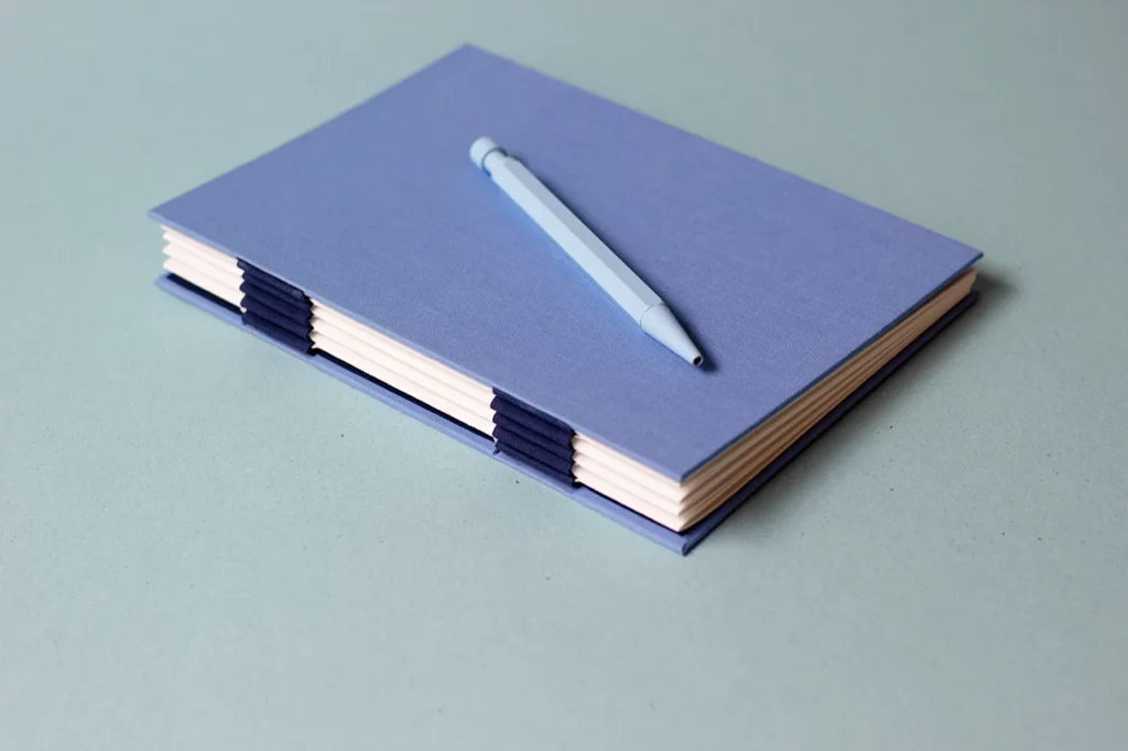 Carnet avec une reliure australienne à charnière piano inversée, style plat, accompagné d’un stylo bleu.