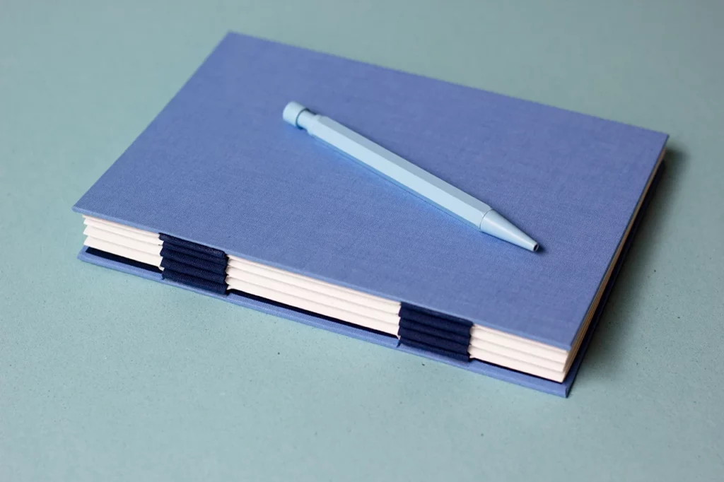 Carnet avec une reliure australienne à charnière piano inversée, style plat, et un stylo bleu (vue 2).