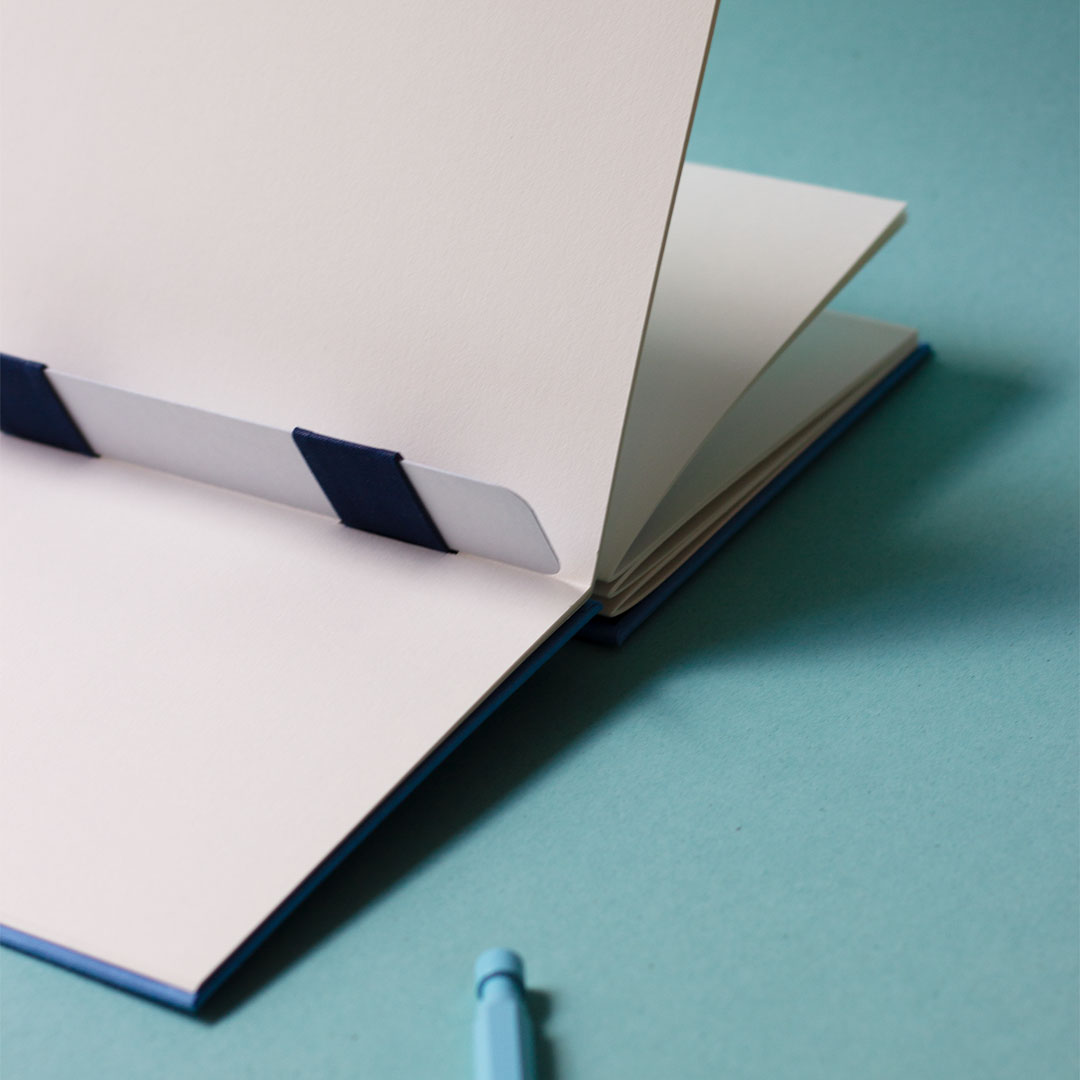 Carnet ouvert avec une reliure australienne à charnière piano inversée, style plat, et un stylo bleu.