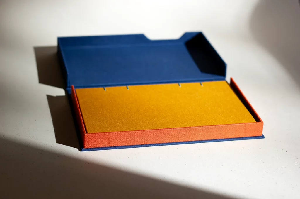 Coffret clamshell ouvert sur mesure, en toile du Marais bleu marine et orange, avec carnet moutarde — Atelier Forme contre forme, Bordeaux — rayon de soleil, vue d’ensemble.