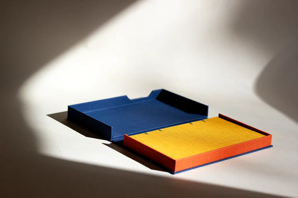 Coffret solander ouvert sur mesure, en toile du Marais bleu marine et orange, avec carnet moutarde, réalisé à l’Atelier Forme contre forme à Bordeaux — rayon de soleil.